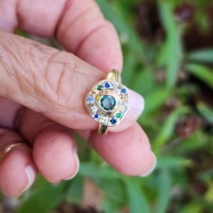 K&ouml;nnte beinhalten: Ein goldener Ring mit einem gr&uuml;nen Edelstein, umgeben von kleineren wei&szlig;en, blauen und gr&uuml;nen Edelsteinen.