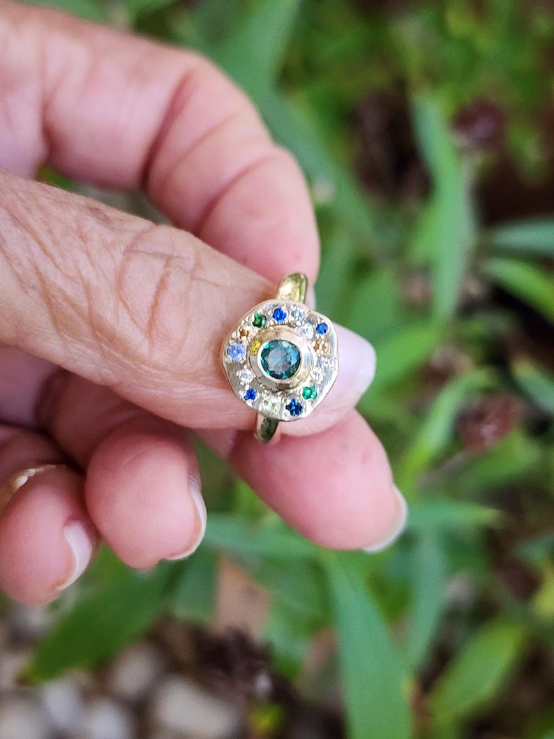 K&ouml;nnte beinhalten: Ein goldener Ring mit einem gr&uuml;nen Edelstein, umgeben von kleineren wei&szlig;en, blauen und gr&uuml;nen Edelsteinen.