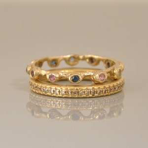 May include: Two gold rings with colorful gemstones. The top ring has a wavy band with pink, blue, and yellow gemstones. The bottom ring has a smooth band with small diamonds.