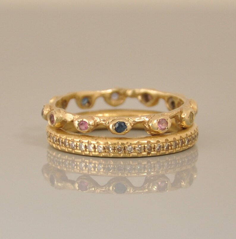 May include: Two gold rings with colorful gemstones. The top ring has a wavy band with pink, blue, and yellow gemstones. The bottom ring has a smooth band with small diamonds.