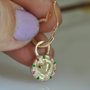 May include: Gold heart-shaped charm with a green and pink gemstone border, attached to a gold hoop earring.