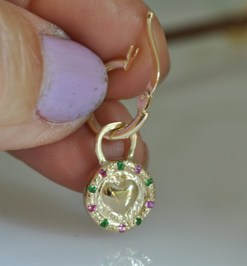 May include: Gold heart-shaped charm with a green and pink gemstone border, attached to a gold hoop earring.