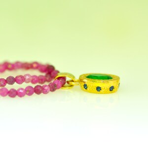 Pu&ograve; includere: Un pendente in oro con una gemma verde al centro e piccole gemme blu. &Egrave; attaccato a una doppia fila di perline rosa. Lo sfondo &egrave; un gradiente da verde chiaro a bianco.
