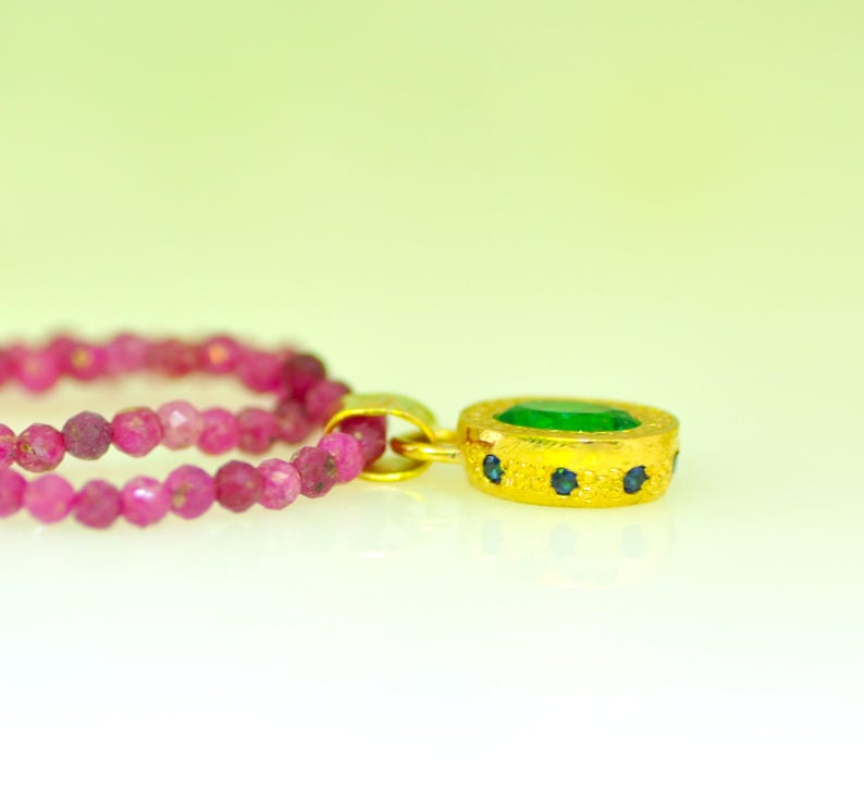 Pu&ograve; includere: Un pendente in oro con una gemma verde al centro e piccole gemme blu. &Egrave; attaccato a una doppia fila di perline rosa. Lo sfondo &egrave; un gradiente da verde chiaro a bianco.