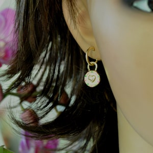 May include: Gold hoop earrings with a small charm dangling from the bottom. The charm is a round disc with a heart design and a border of colorful gemstones.