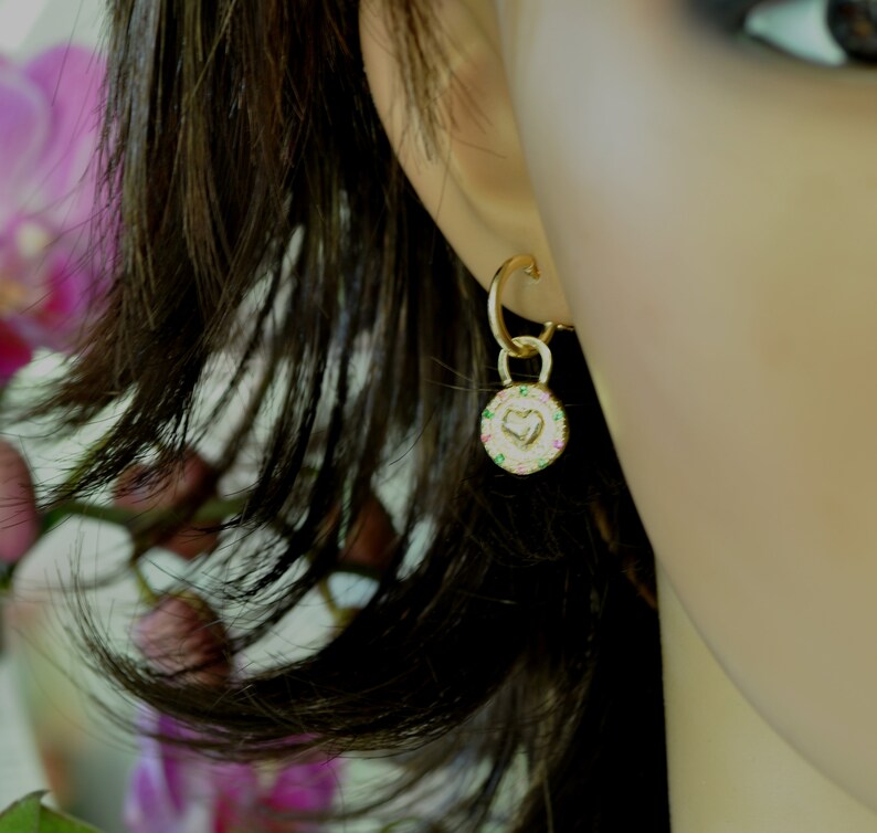 May include: Gold hoop earrings with a small charm dangling from the bottom. The charm is a round disc with a heart design and a border of colorful gemstones.