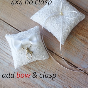 May include: Two white lace ring pillows. The smaller pillow is 3x3 inches and has a white satin bow and a gold clasp. The larger pillow is 4x4 inches and has a white satin ribbon with a pearl.