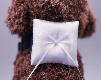 May include: A brown plush dog wearing a white pillow with a white satin bow and a pearl in the center. The pillow is attached to the dog with a white satin ribbon.