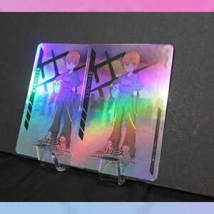 May include: Two holographic trading cards featuring an anime-style character with orange hair, wearing a purple shirt and green pants. The cards have Japanese text and the word "RESOURCE" in green.