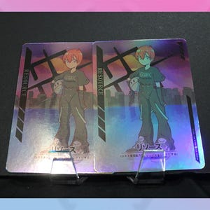 May include: Two holographic trading cards featuring a cartoon character with red hair and a green outfit. The cards have the word "RESOURCE" printed vertically on the left side. The background has a cityscape design.