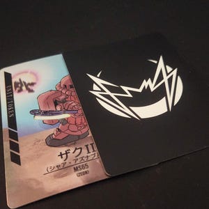 Puede incluir: Dos cartas coleccionables. Una carta presenta un robot rojo con un arma, el texto "ザク II" y "MS65 (ZEON)". La otra carta es negra con un dise&ntilde;o gr&aacute;fico blanco. Las cartas est&aacute;n sobre una superficie negra.