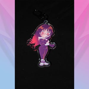 May include: A purple anime-style keychain featuring a character with red hair and eyes, wearing a purple outfit. The keychain has a black clasp and is set against a black background with a pink and blue gradient.