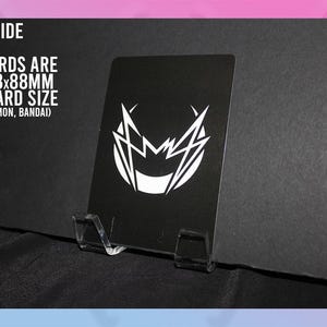 Puede incluir: Una tarjeta negra con un dise&ntilde;o gr&aacute;fico blanco, exhibida en un soporte de pl&aacute;stico transparente. Se ve el reverso de la tarjeta, con texto que indica un tama&ntilde;o de 63x88mm. El texto tambi&eacute;n menciona MTG, Pokemon y Bandai.