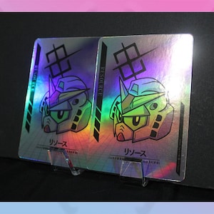 May include: Two holographic trading cards featuring a robot head design with green and yellow accents. The cards have Japanese text and the word "RESOURCE" printed on the side. They are displayed on a clear stand.