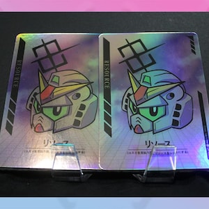 May include: Two holographic trading cards featuring a robot head design with green eyes, a yellow crest, and the word "RESOURCE" in a vertical column. Japanese text is also present. The cards are displayed on clear stands.