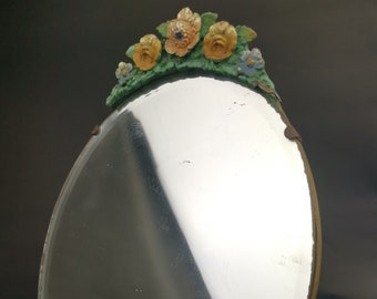 Puede incluir: Un espejo de forma ovalada con una cresta decorativa superior adornada con flores esculpidas en tonos amarillos y naranjas entre hojas verdes. El cristal del espejo se sujeta con pequeños clips metálicos en el borde, mostrando una superficie reflectante.