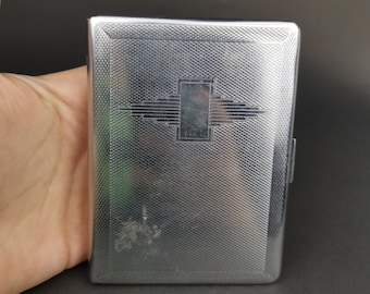 May include: A rectangular silver-toned metal cigarette case featuring a fine cross-hatched texture across the surface. The center of the case displays a smooth, polished rectangular emblem with horizontal stepped lines extending outward on both sides.