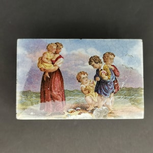 May include: A rectangular ceramic box lid featuring a painted scene of a person holding a child in a yellow dress, standing near three other children on a beach. The background shows a blue ocean, a cloudy sky, and two flying seagulls.
