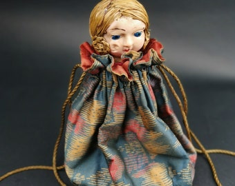 May include: A drawstring pouch featuring a doll head with blonde hair and blue eyes. The fabric is a teal, red, and gold patterned textile with a ruffled collar. Two thin gold cords serve as the closure mechanism for the bag.