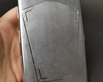 May include: A rectangular silver-toned metal cigarette case featuring a fine cross-hatched texture across the surface. The front panel displays a central shield-like shape outlined with engraved decorative lines and corner accents.