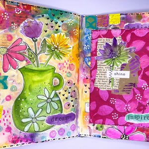 May include: A vibrant, open journal spread with colorful mixed-media art. A green vase with white flowers holds pink, purple, and yellow flowers. The word "dream" is visible. The right page features a pink floral design with the words "shine" and "inspire".