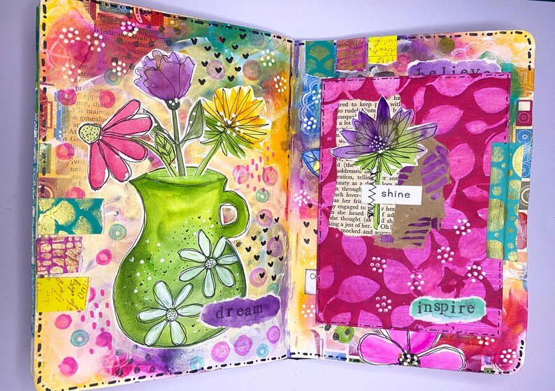 May include: A vibrant, open journal spread with colorful mixed-media art. A green vase with white flowers holds pink, purple, and yellow flowers. The word "dream" is visible. The right page features a pink floral design with the words "shine" and "inspire".