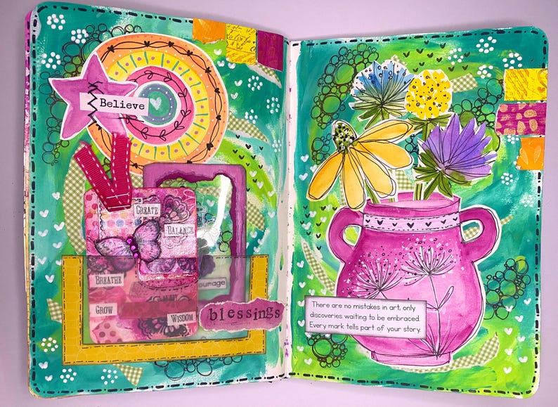 May include: A vibrant, open journal spread with mixed media art. The left page features a star with "Believe" and a collage with "Create, Balance, Breathe, Grow, Wisdom, Blessings". The right page has a pink vase with flowers and text: "There are no mistakes in art..."