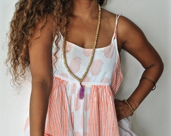 May include: A woman wearing a white and orange patterned dress with thin straps. The dress features a paisley print on the bodice and vertical stripes on the skirt. A long wooden bead necklace with a purple tassel hangs down.