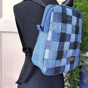 May include: A denim backpack with a woven design in shades of blue and black. The backpack has a zippered main compartment, a small front pocket, and adjustable black straps. A small, round, bronze-colored emblem is on the front.