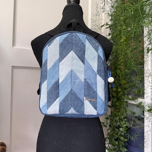 May include: A denim backpack with a chevron pattern in shades of blue and white. The backpack is displayed on a black mannequin. The bag has a small, round charm attached to the zipper. The brand name is visible on the front of the bag.