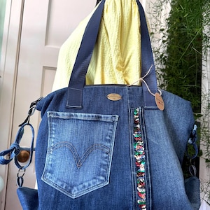 May include: A denim tote bag with a large front pocket and a decorative sequin stripe. The bag features a dark blue strap and a small tag. The denim has a faded wash, and the bag appears to be handmade.