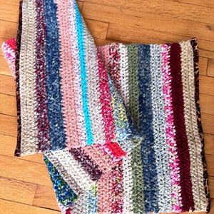 May include: A handmade, colorful, striped blanket with a variety of colors including pink, blue, and green. The blanket has a textured appearance and a leopard print border. The blanket is laying on a wooden floor.