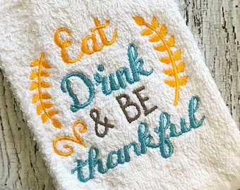 May include: White kitchen towel with embroidered text that reads "Eat Drink & Be Thankful" in teal, brown, and yellow lettering. The text is surrounded by yellow embroidered leaves.