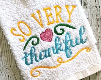 May include: White kitchen towel with embroidered text "So very thankful" in yellow, blue, and green with a pink heart in the center.