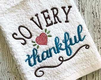 May include: White kitchen towel with brown embroidery that reads "So very thankful" with a red heart and green leaves.