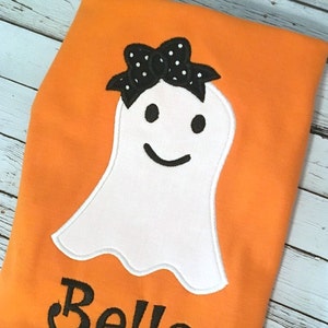 Puede incluir: Tela naranja con un fantasma blanco con un lazo negro y ojos negros y una sonrisa negra. El texto "Bella" est&aacute; debajo del fantasma.