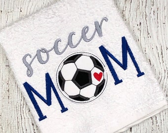 May include: White towel with the text "soccer MOM" embroidered in blue and gray. A black and white soccer ball with a red heart is embroidered in the center of the towel.