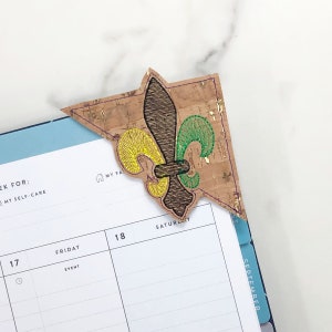 May include: A cork bookmark with a green, yellow, and brown embroidered fleur-de-lis design. The bookmark is shaped like a corner and is placed on a page of a planner.