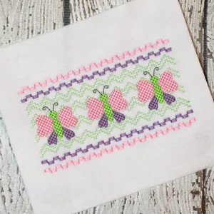 May include: A white fabric with a pink, green, and purple embroidered border featuring three butterflies. The butterflies have pink wings with green and purple accents.