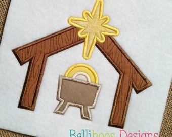 May include: Embroidered design of a wooden manger with a star above it. The manger is brown and the star is yellow. The text "Stitched by Shelly Christensen" and "Belliboos Designs" are also visible.