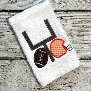May include: White towel with an embroidered football, football helmet, and goal post. The football is brown with white laces. The helmet is orange with a white stripe. The goal post is gray.