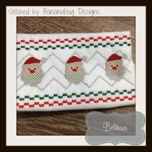 Puede incluir: Tela blanca con un borde rojo y verde y tres caras de Papá Noel bordadas. La tela tiene un patrón en zigzag y el texto "Stitched by: Bananabug Designs" está en la parte superior.