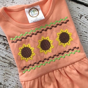 Pode incluir: Um vestido sem mangas cor pêssego com um padrão bordado de girassóis marrons e verdes. O vestido tem um decote redondo e uma saia franzida. A etiqueta do vestido diz "BB Bella Baby" e "3T 100% algodão penteado Feito na China."