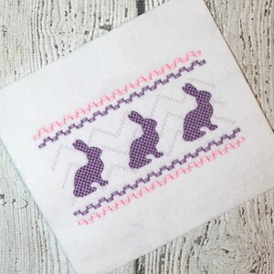 May include: Purple embroidered bunny silhouettes on a white fabric with pink and purple zig-zag stitching.