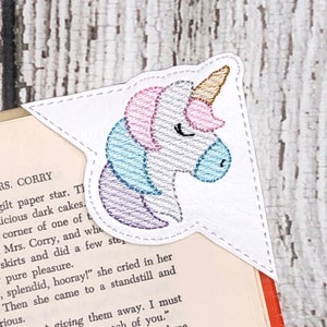 Könnte beinhalten: Ein weißes Einhorn-Lesezeichen in Form eines Dreiecks mit abgerundeten Ecken. Das Lesezeichen ist mit einem Einhornkopf bestickt. Die Mähne und das Horn des Einhorns sind rosa und blau.