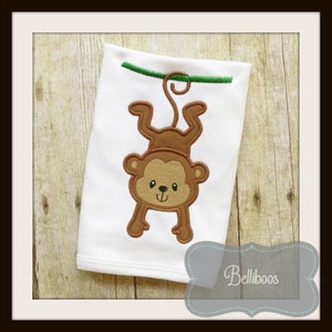 May include: A white babygrow with an embroidered brown monkey hanging upside down from a green vine. The monkey has a brown face with black eyes and a pink nose.