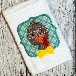 May include: A white towel with an embroidered turkey wearing glasses and a yellow bow tie. The turkey is surrounded by a green and white checked frame.