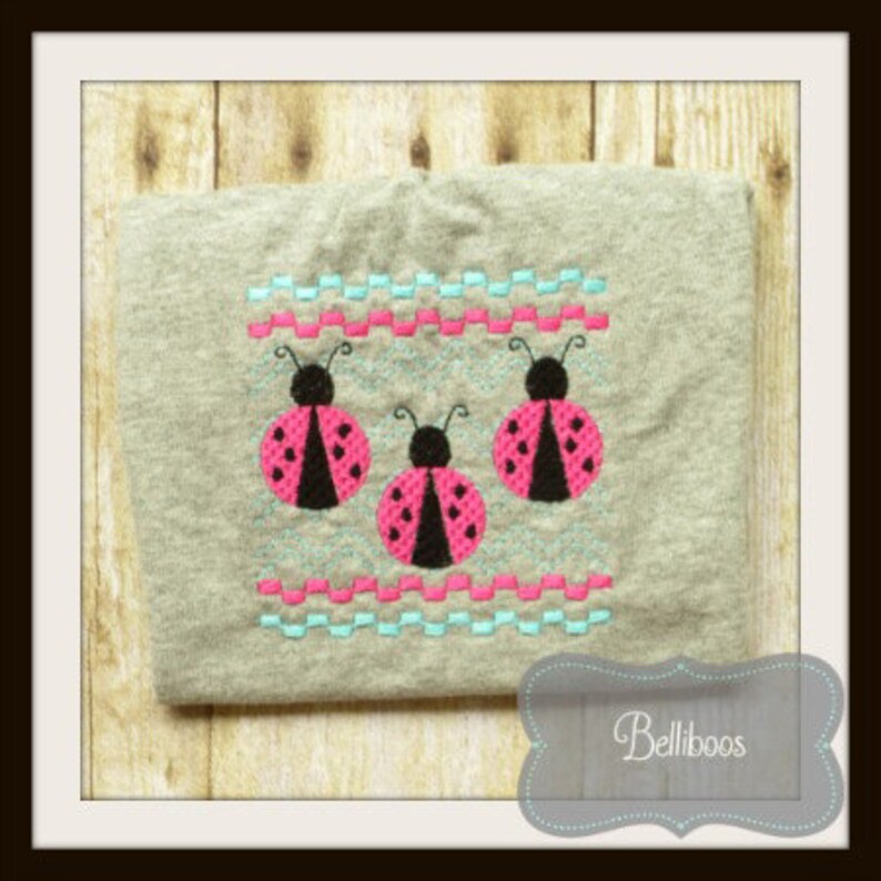 May include: A gray t-shirt with three pink and black ladybugs embroidered on it. The ladybugs are arranged in a row and are surrounded by a blue and pink zig-zag pattern.