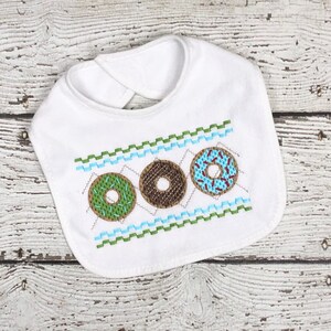 May include: White baby bib with a green, brown, and blue embroidered donut design. The donuts have sprinkles and a hole in the center.