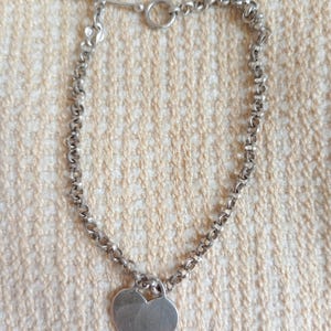 May include: A silver-toned necklace with a heart-shaped pendant. The necklace features a chain with a toggle clasp closure. The heart pendant is smooth and polished. The necklace is displayed on a textured, light-colored surface.
